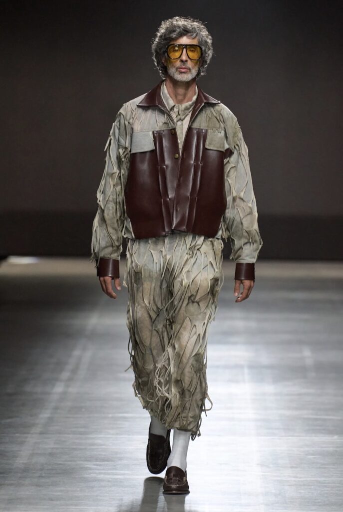 Male model walking down runway wearing textured beige outfit with brown leather details. 