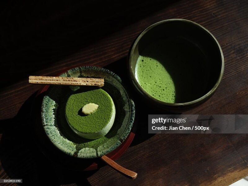 High view angle of a round matcha cake next to a cup of hot matcha tea on a dark wooden table.