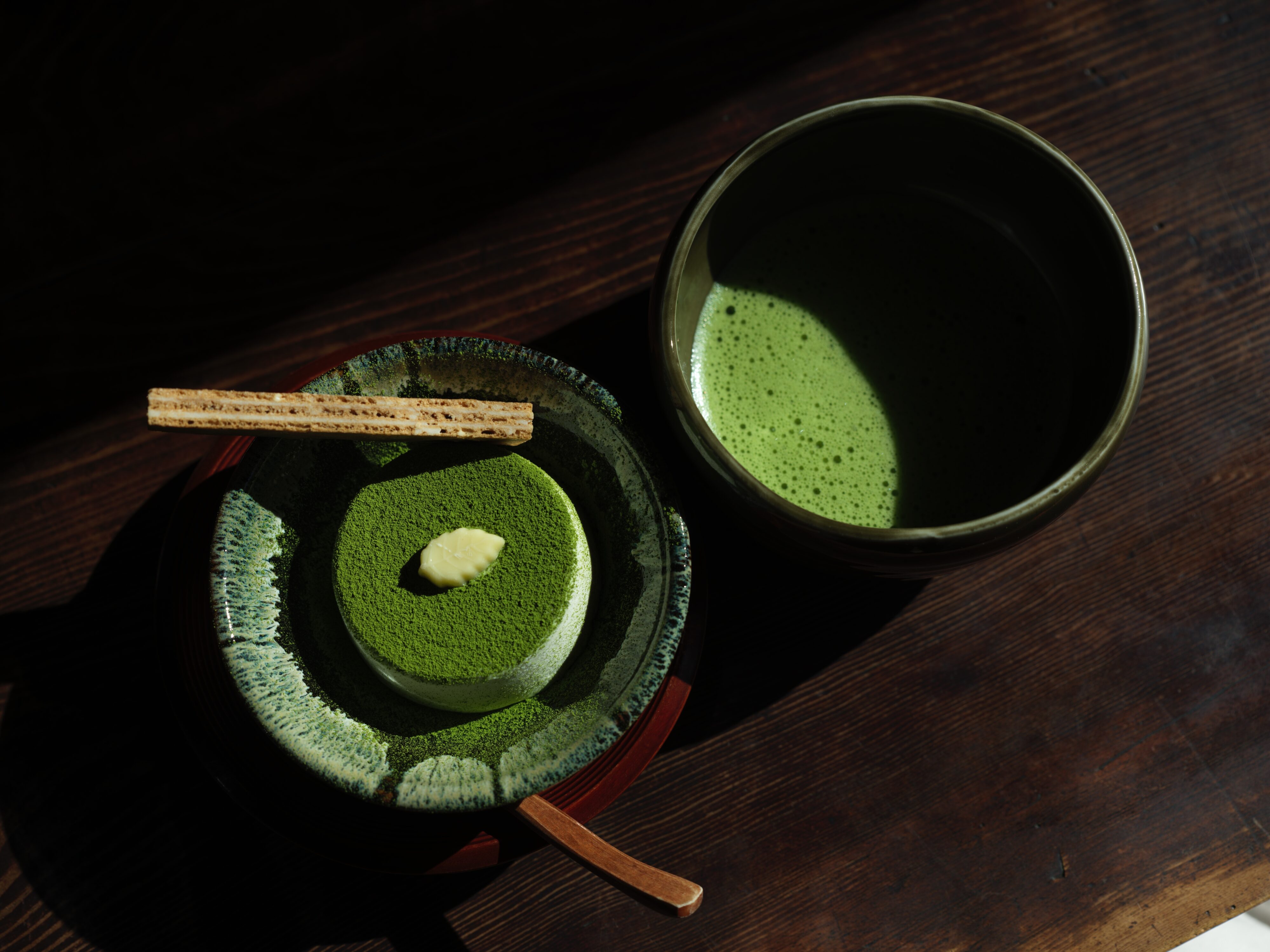 High view angle of a round matcha cake next to a cup of hot matcha tea on a dark wooden table.