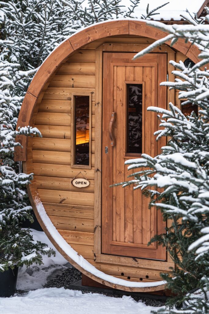 Round wooden sauna surrounded by snowy trees. 