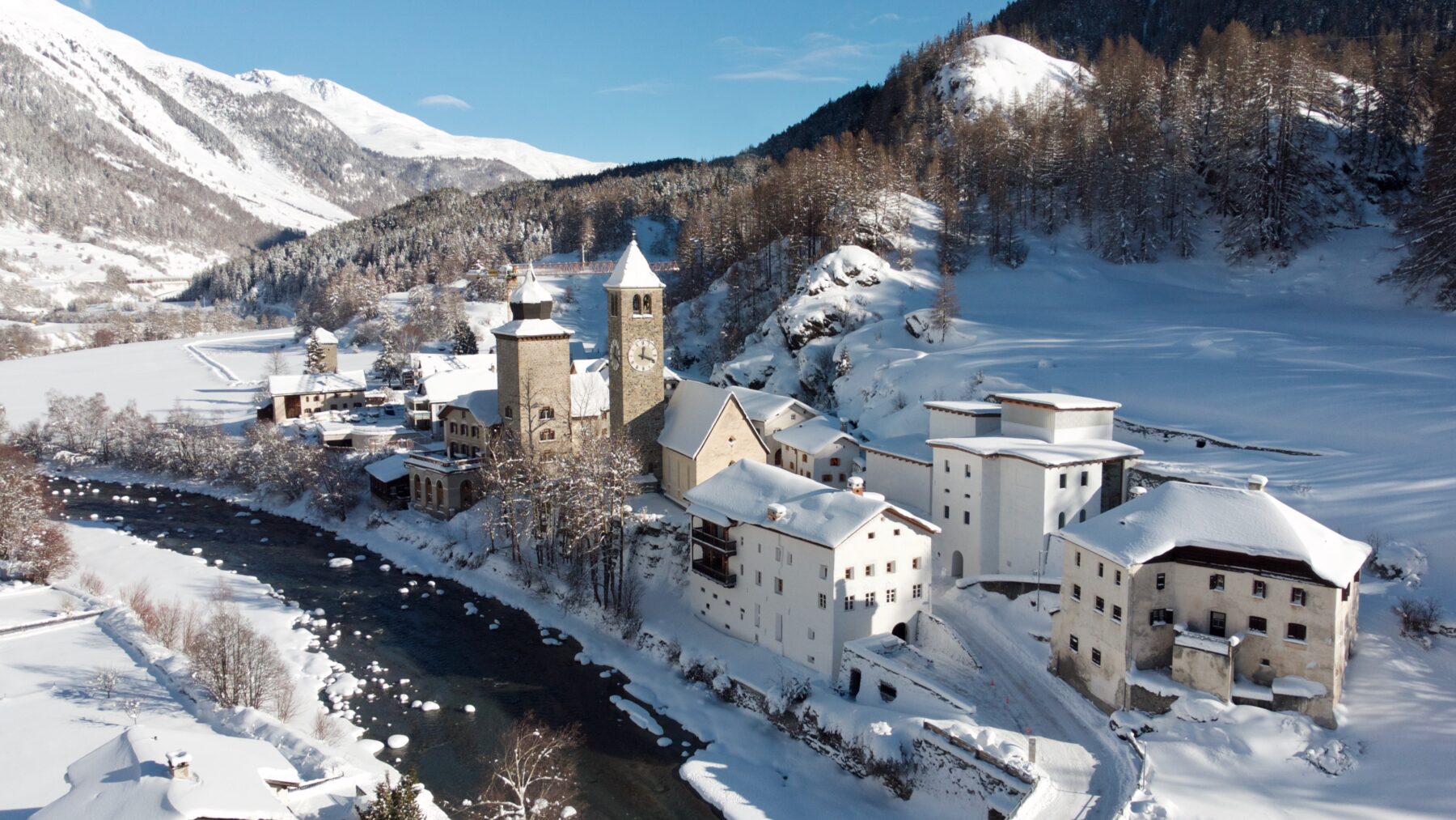 An alpine village covered in snow with a flowing river in the middle and mountains in the background.