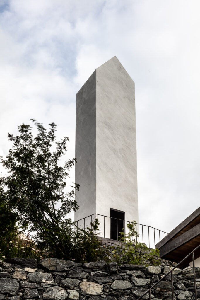 Tall beige stone tower with a slanted, triangular roof surrounded by stones and greenery.