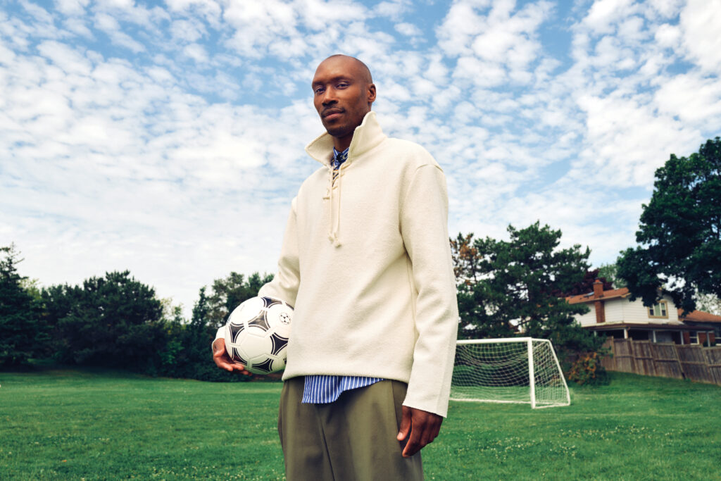 Atiba Hutchinson standing on a soccer field holding a soccer ball, wearing a cream-colored sweater and blue striped shirt, with a goalpost and trees in the background, reflecting on his multicultural identity and soccer career.