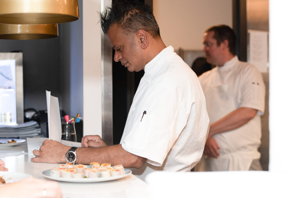Chef Sash Simpson reviewing dishes in a restaurant kitchen, with sushi on a plate and a fellow chef in the background, highlighting his culinary expertise and dedication to his craft.