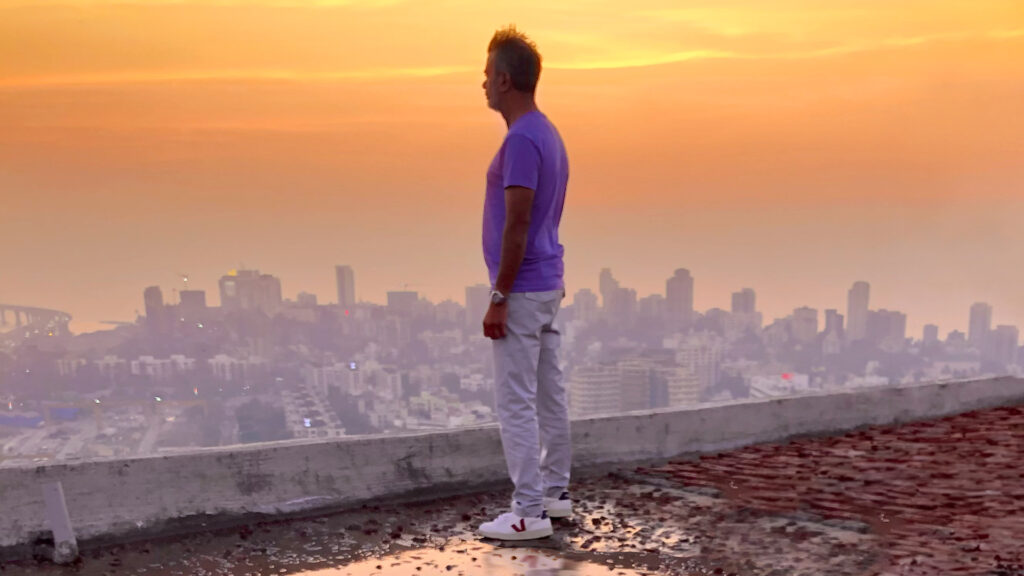 Man standing on a rooftop overlooking a hazy city skyline at sunset, reflecting on his journey as a celebrated chef and philanthropist.