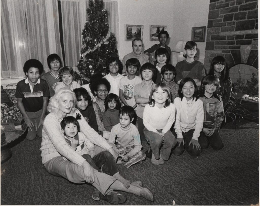 Group of children and adults gathered around a Christmas tree, highlighting community and family connections, reflecting Sash Simpson's philanthropic focus on orphans and childhood experiences.