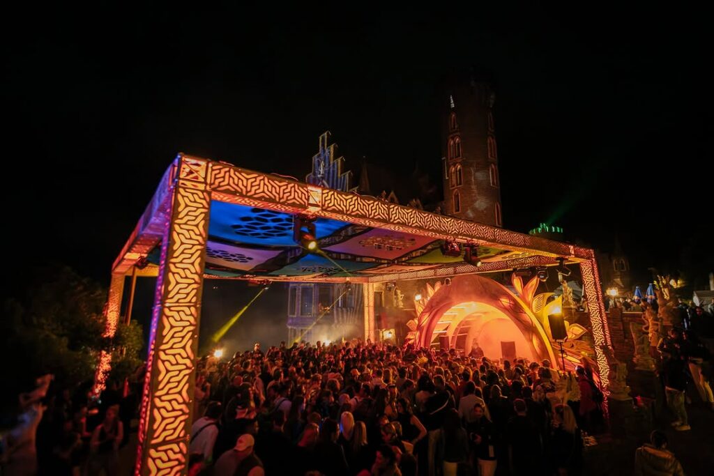 Crowd celebrating New Year's Eve at a vibrant outdoor festival stage illuminated with colorful lights, featuring artistic designs and a historic castle backdrop.