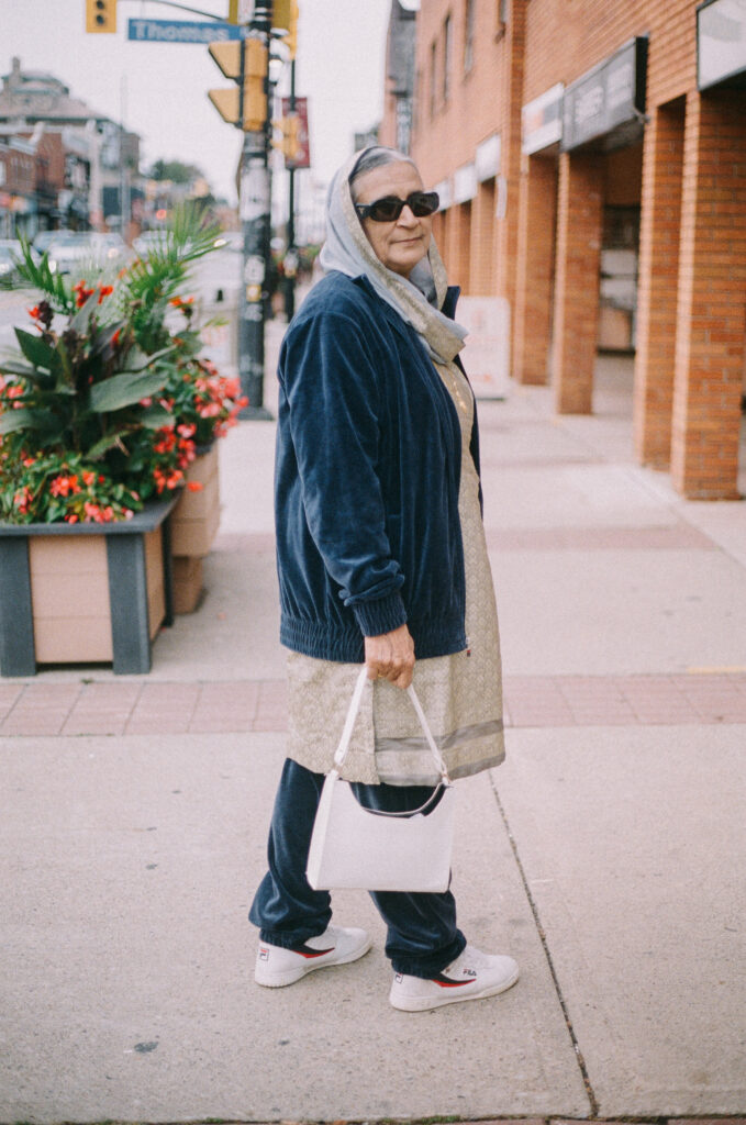 Stylish senior woman in casual outfit, wearing sunglasses and a headscarf, walking on city street, embodying Grandmacore fashion trend.