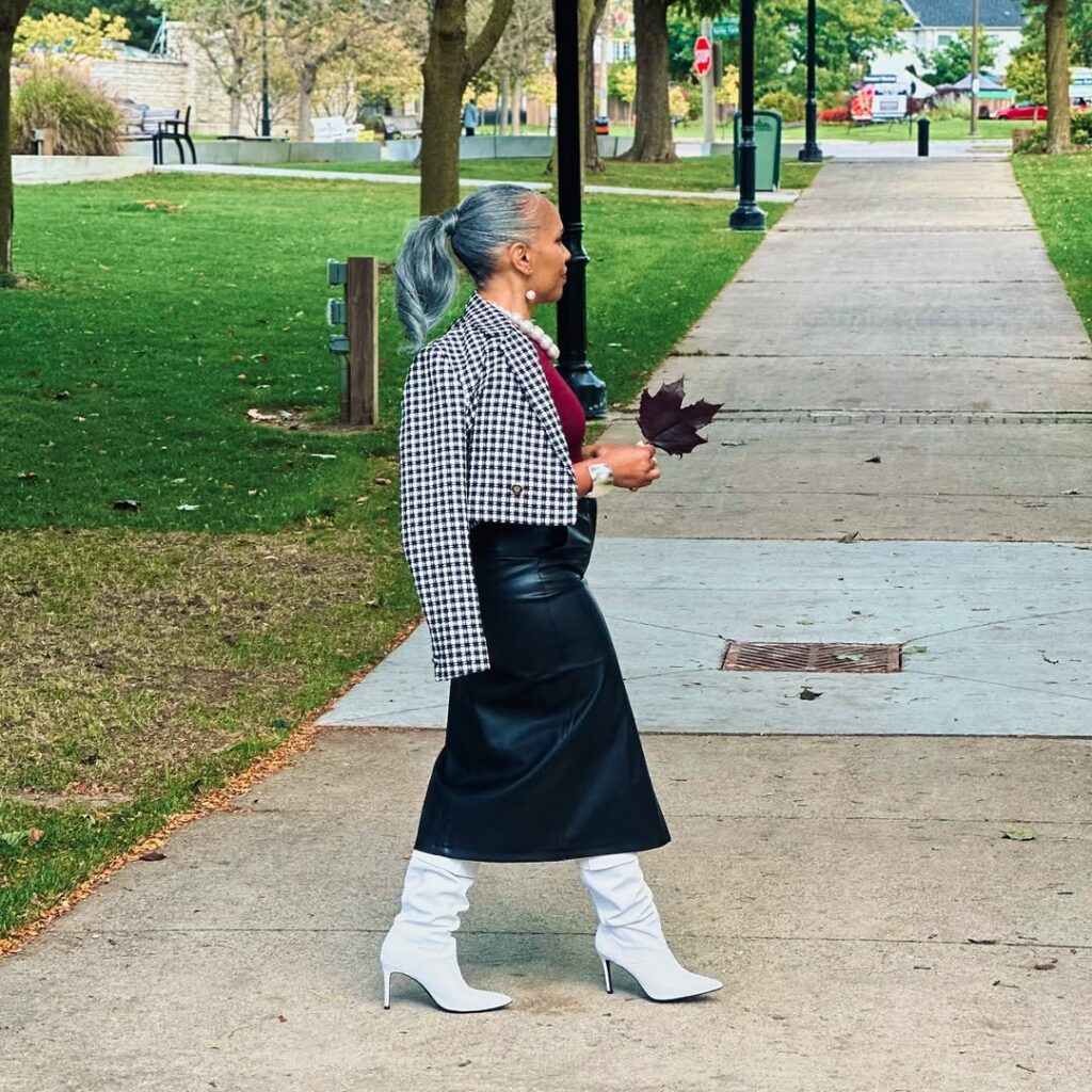 Stylish senior woman walking in a park, wearing a checkered blazer, a maroon top, a black leather skirt, and white high-heeled boots, holding a red leaf, embodying the Grandmacore fashion trend.