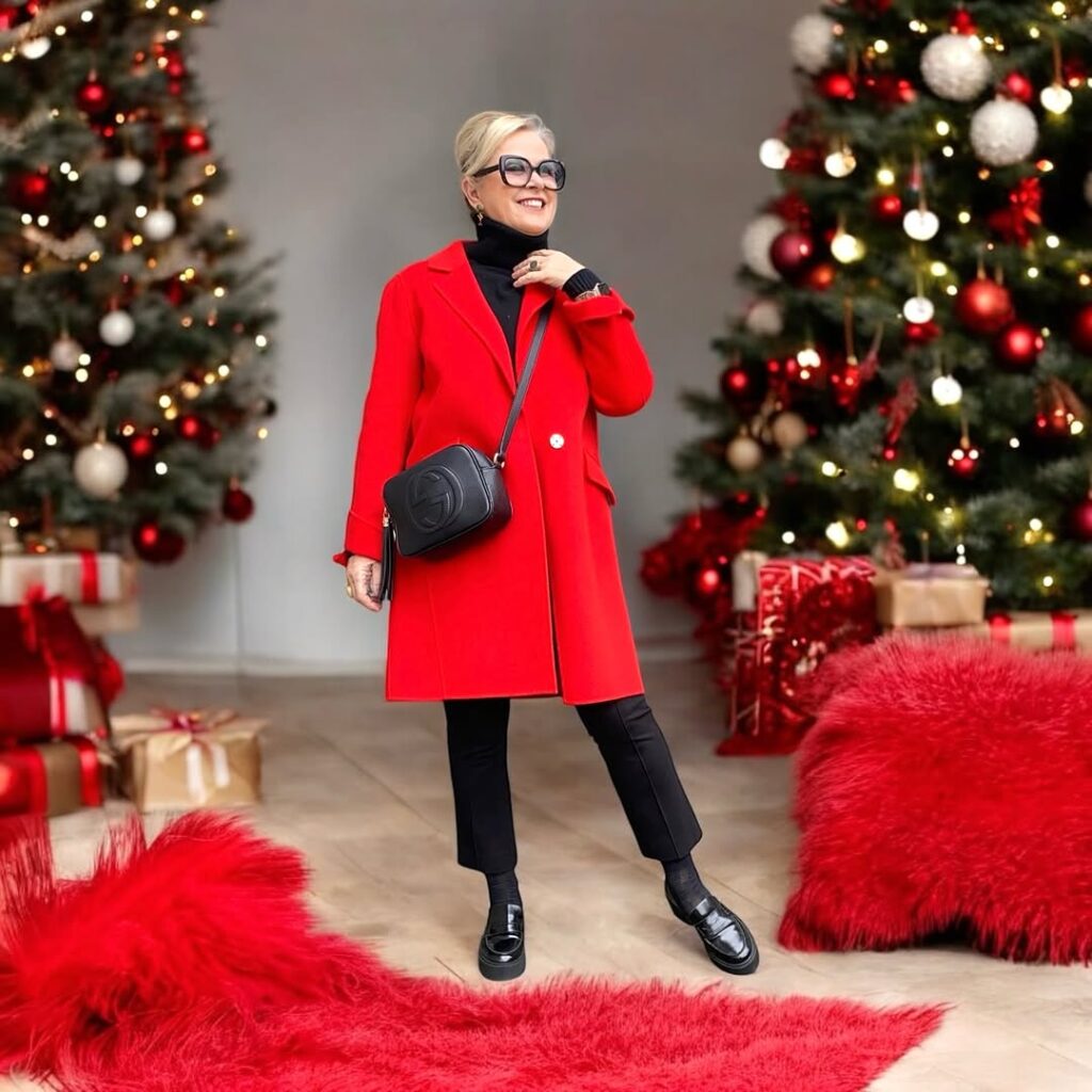 Stylish senior woman in a vibrant red coat and black pants, posing in front of a decorated Christmas tree, embodying the Grandmacore fashion trend.