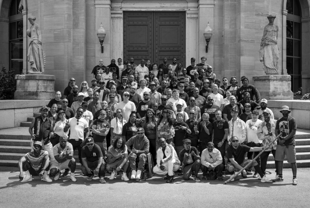 Group of diverse individuals gathered on the steps of the Art Gallery of Ontario, celebrating hip-hop culture, with a statue in the background, representing community and artistic expression.