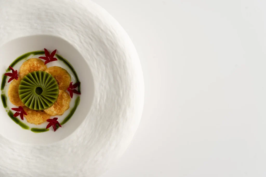 Artfully presented South Asian fine dining dish featuring green mousse, golden discs, and edible flowers on a textured white plate, emphasizing culinary innovation.