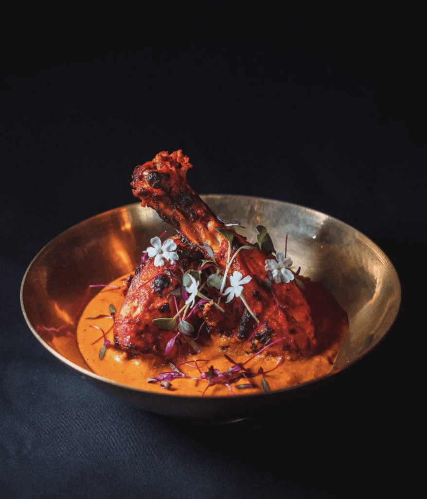 Grilled chicken drumsticks in a rich, creamy sauce, garnished with edible flowers and microgreens, presented in a gold bowl on a dark background, highlighting South Asian fine dining cuisine.