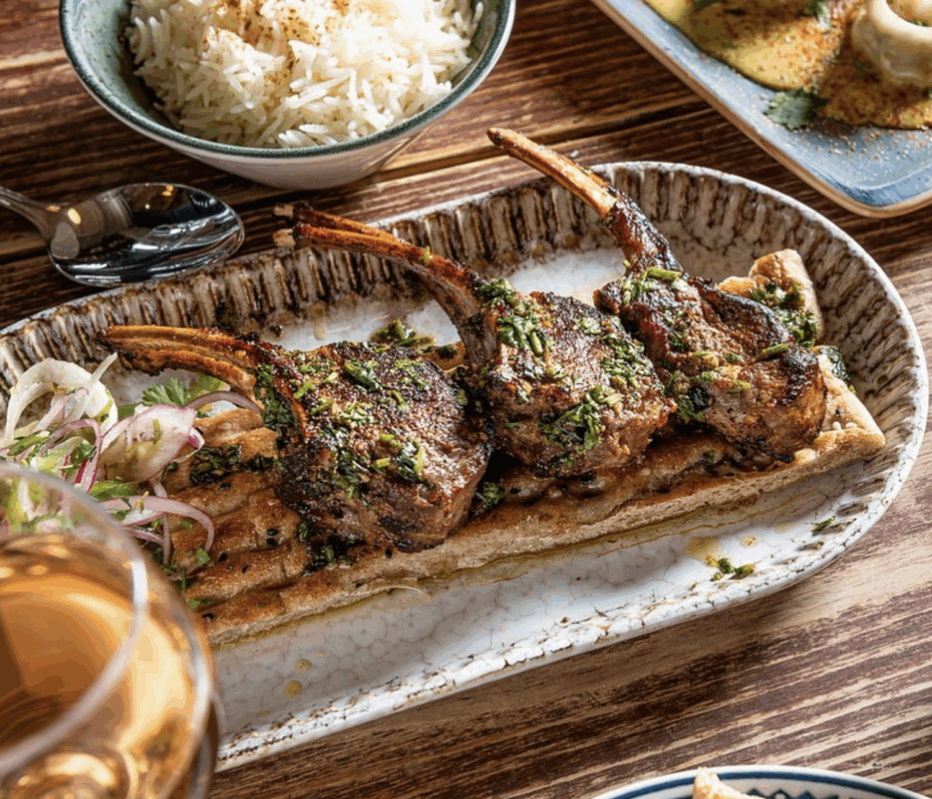 Lamb chops garnished with herbs on a platter, accompanied by rice and side salad, showcasing South Asian fine dining cuisine.