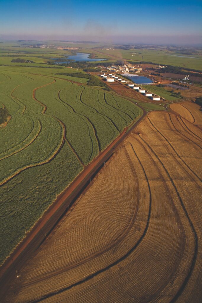 Aerial view of agricultural fields with wavy patterns, oil storage tanks, and a river, illustrating the intersection of energy production and land use in emerging markets like Guyana.